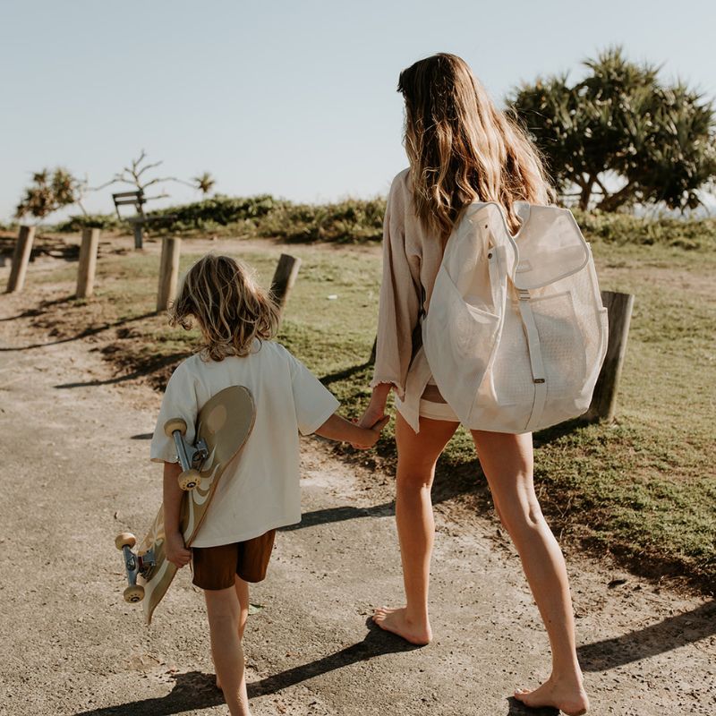 Mum walking to the beach with her son, carrying the Sande Kids Beach Hauler Mesh Backpack in neutral Sand, showcasing its spacious design and durable construction for carrying family beach essentials.