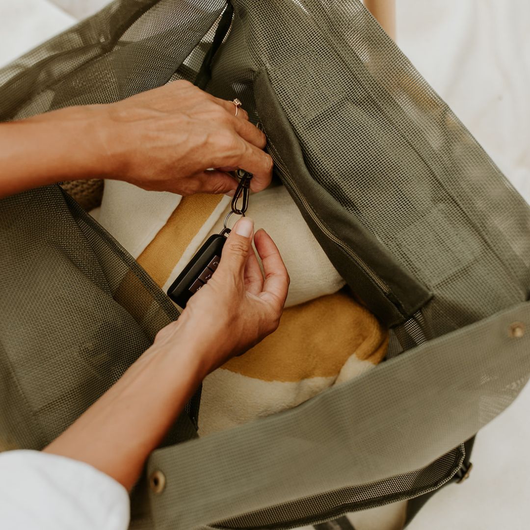 The top view of the Sande Kids™ Beach Hauler mesh backpack in Pandanus green, filled with beach towels. A woman’s hands are seen clipping car keys to the internal key clip inside the secure valuables pocket.