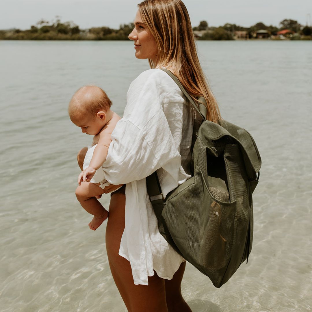 A mum with long blonde hair stands on the side at the water’s edge, showcasing the side view of the dark olive green Sande Kids™ Beach Hauler mesh backpack. She’s holding a newborn baby in her arms. The backpack features side pockets, a flap with a clip, and a white silicone logo, making it both stylish and functional for family outings at the beach.