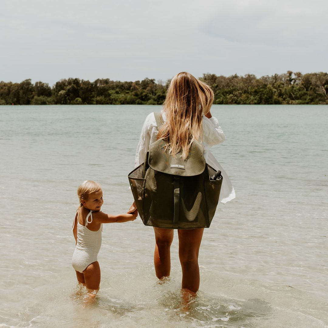 A woman with long blonde hair stands at the water’s edge, holding her little girl’s hand. She wears a dark olive green Sande Kids™ Beach Hauler mesh backpack, which features side pockets, a flap with a clip, and a white silicone logo. The scene captures a peaceful beach moment, highlighting the backpack’s style and practicality for family outings.