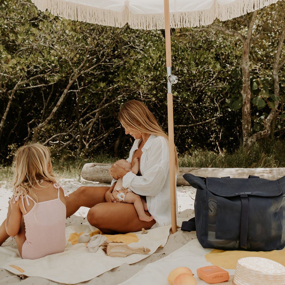A mum sits on a towel under a beach umbrella, breastfeeding her newborn while chatting with her little girl. Next to them is the Sande Kids™ Beach Hauler mesh backpack in Deep Sea Navy.