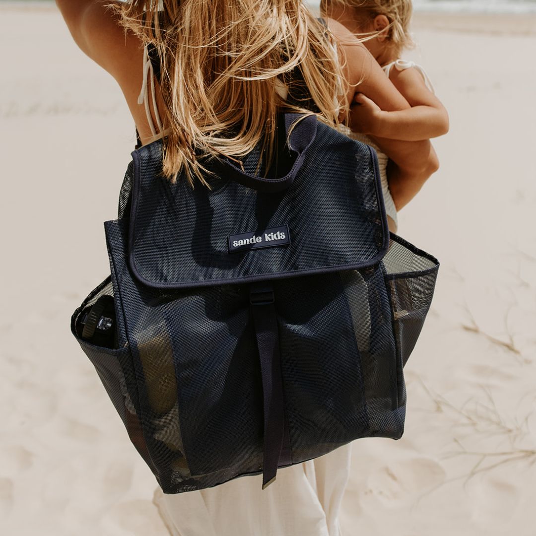 A mum is walking along the beach wearing the Sande Kids™ Deep Sea Navy Beach Hauler mesh backpack, demonstrating hands-free carrying as she has a toddler on her hip.