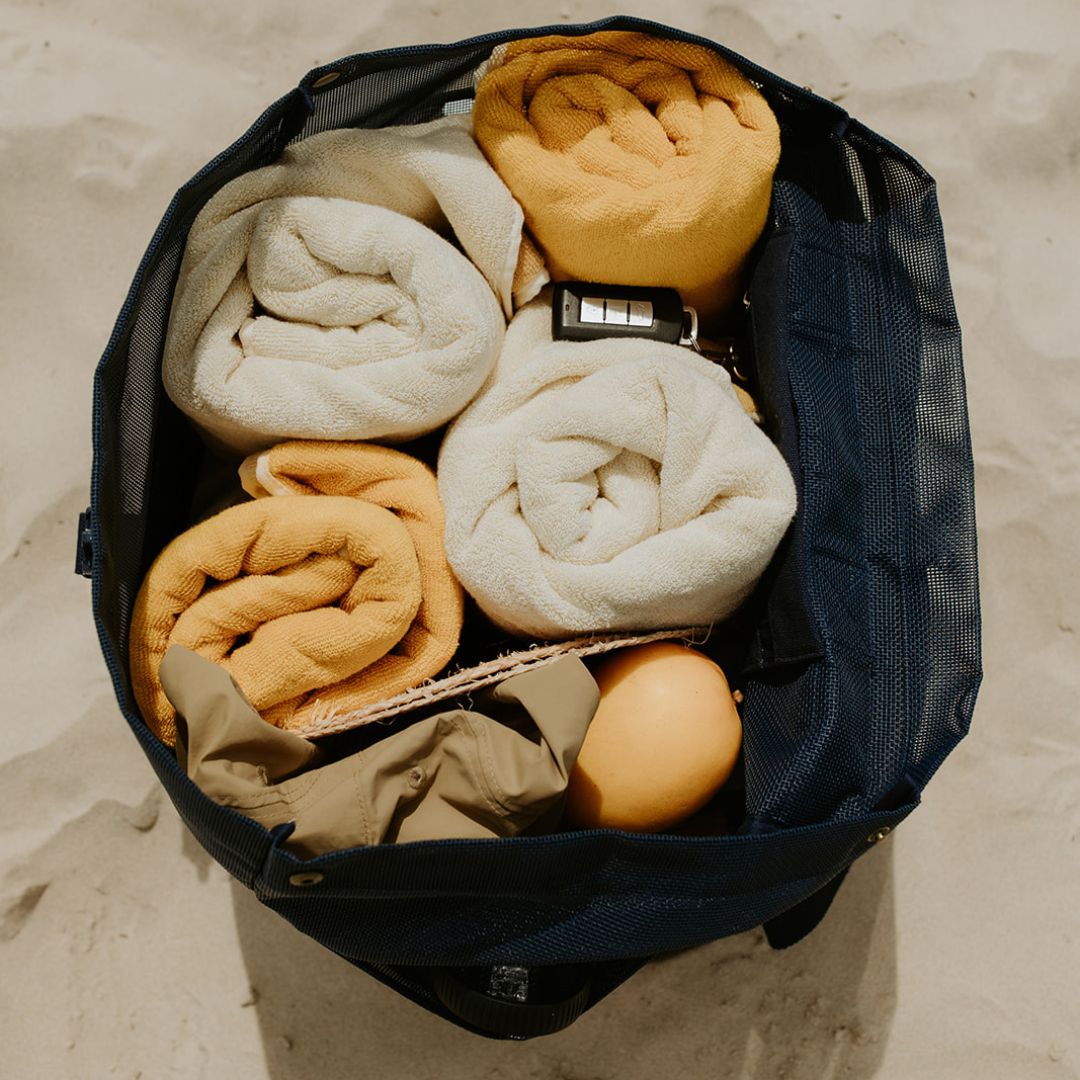 The Sande Kids™ Beach Hauler mesh backpack in Deep Sea Navy sits on the sand, packed with four bulky beach towels, a hat, sunscreen, snacks, and valuables, demonstrating its large capacity.