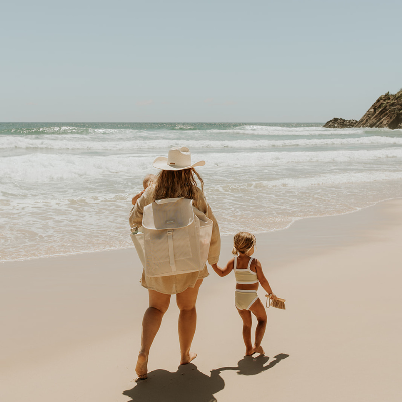 A woman and young girl walk hand in hand along the beach. The woman carries a cream Sande™ Beach Hauler Mesh Backpack and wears a straw hat and beige cover-up.