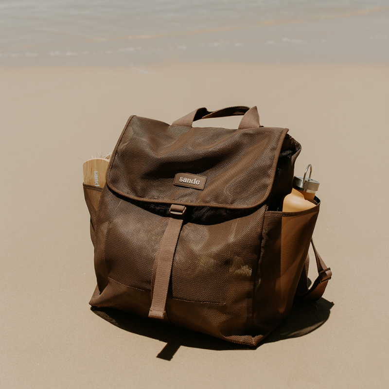 Beach lifestyle shot featuring the Sande Beach Hauler Mesh Backpack in Espresso chocolate brown, highlighting its durable construction and spacious design for family beach adventures. Backpack is sitting the sand.