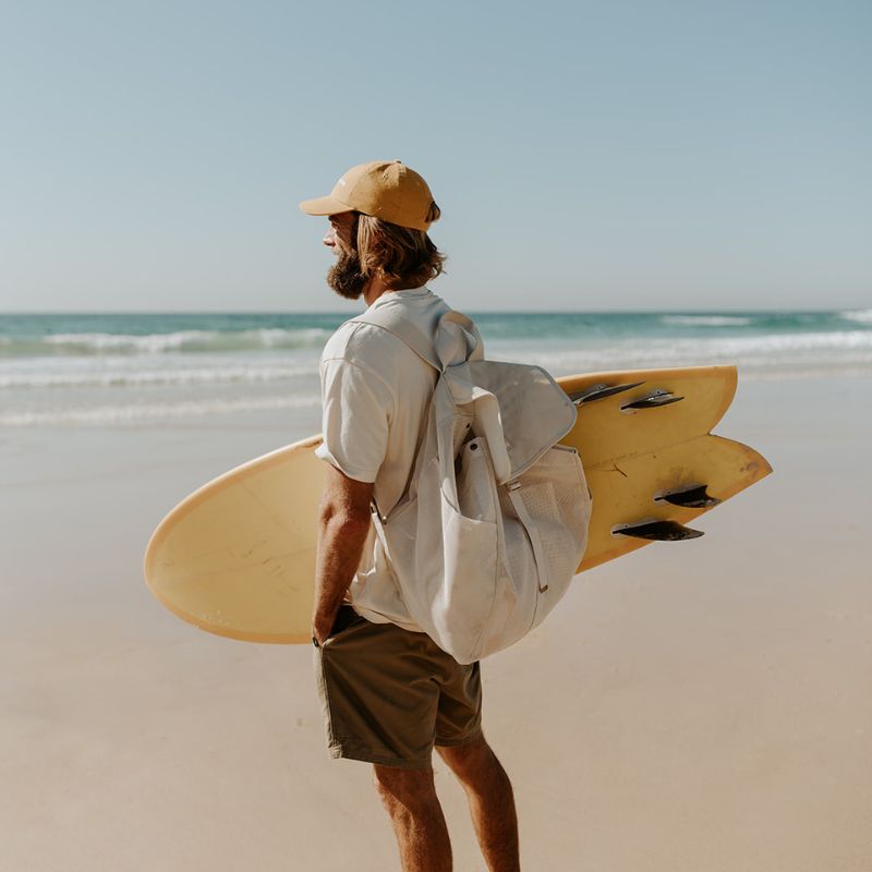 Dad at the beach carrying a yellow surfboard under one arm and wearing the Sande Kids Beach Hauler Mesh Backpack on his back, showcasing its spacious, durable design for carrying beach essentials on family and surf outings.