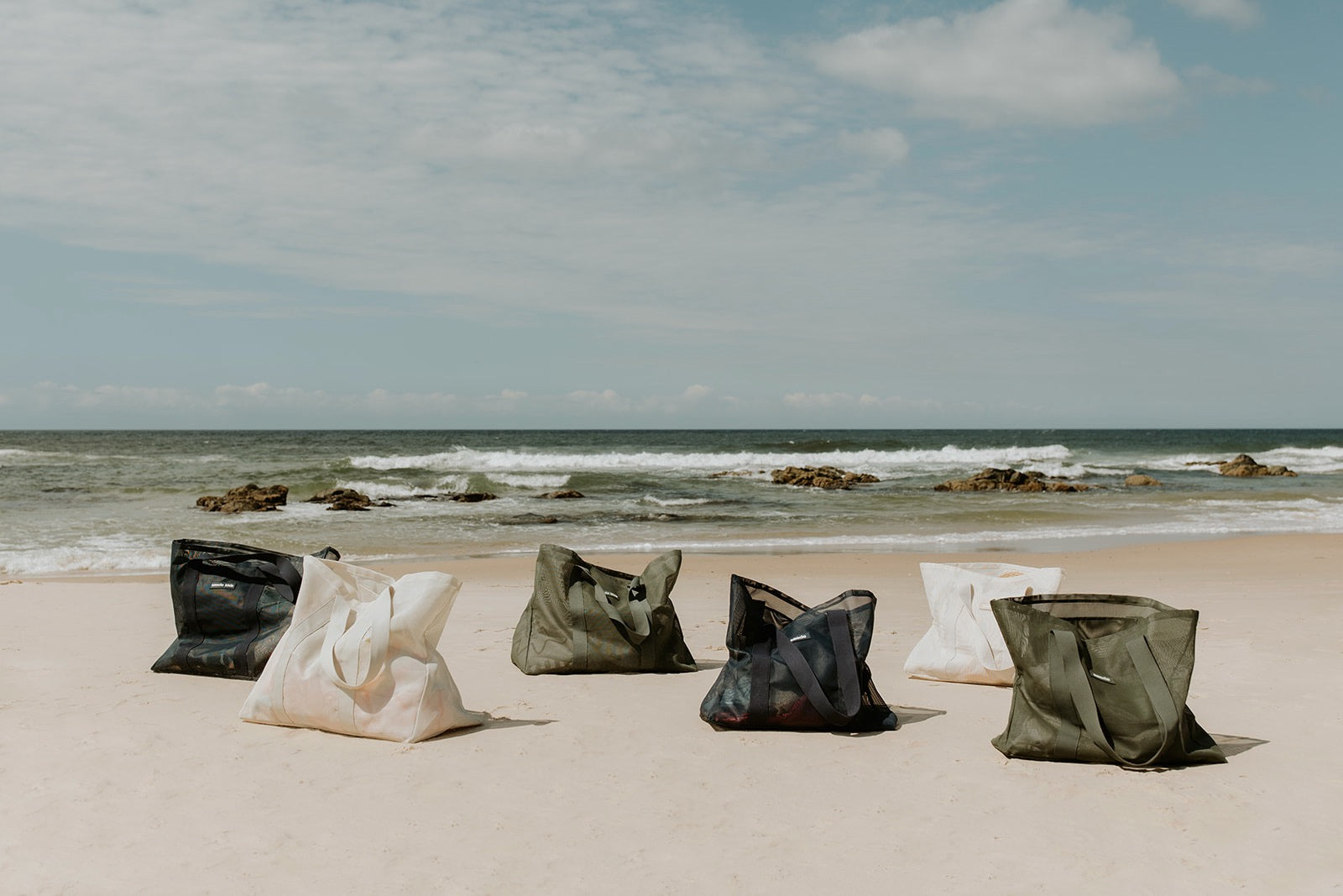 The Sande Kids Beach Hauler mesh beach bag range - oversized Beach Hauler and Beach Hauler - Mini in neutral Sand, Pandanus green and Deep Sea navy. Lined up on the sand with ocean and sky in the background.