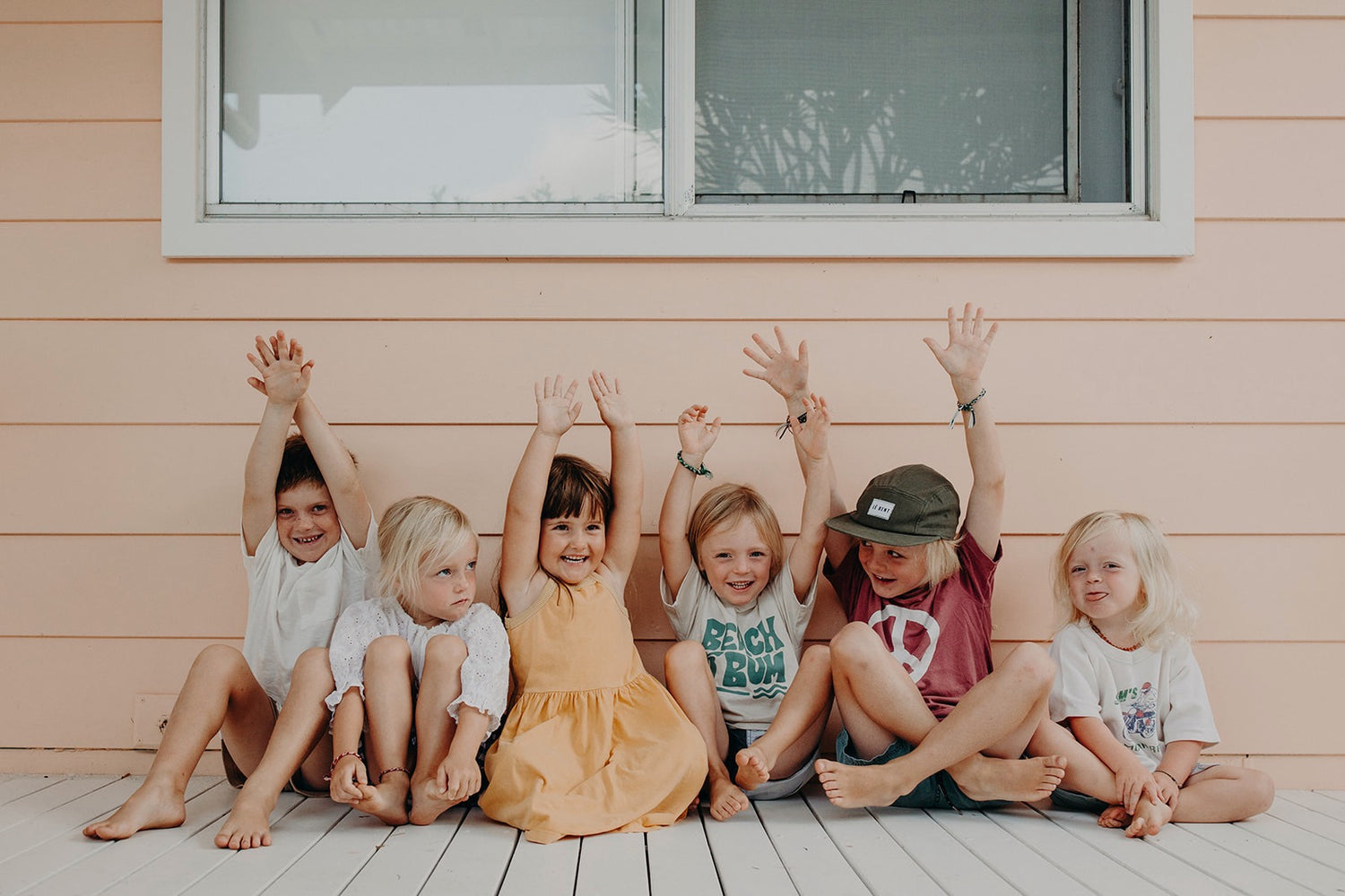 Six little kids, 3 years to 6 years sitting on a white deck in front of a peach weatherboard house. The About Us image for Sande Kids.