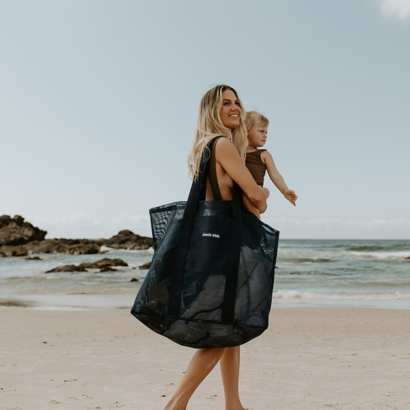 Oversized mesh beach bag, the Sande Kids Beach Hauler in Deep Sea Navy, carried by a mum holding her young daughter at the beach, with the ocean in the background.