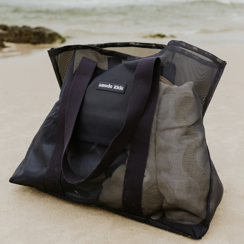 A close-up image of the Sande Kids Beach Hauler mesh beach bag in Deep Sea Navy, packed with towels and beach day essentials. The bag sits on the sand, with the ocean and rocks softly blurred in the background.