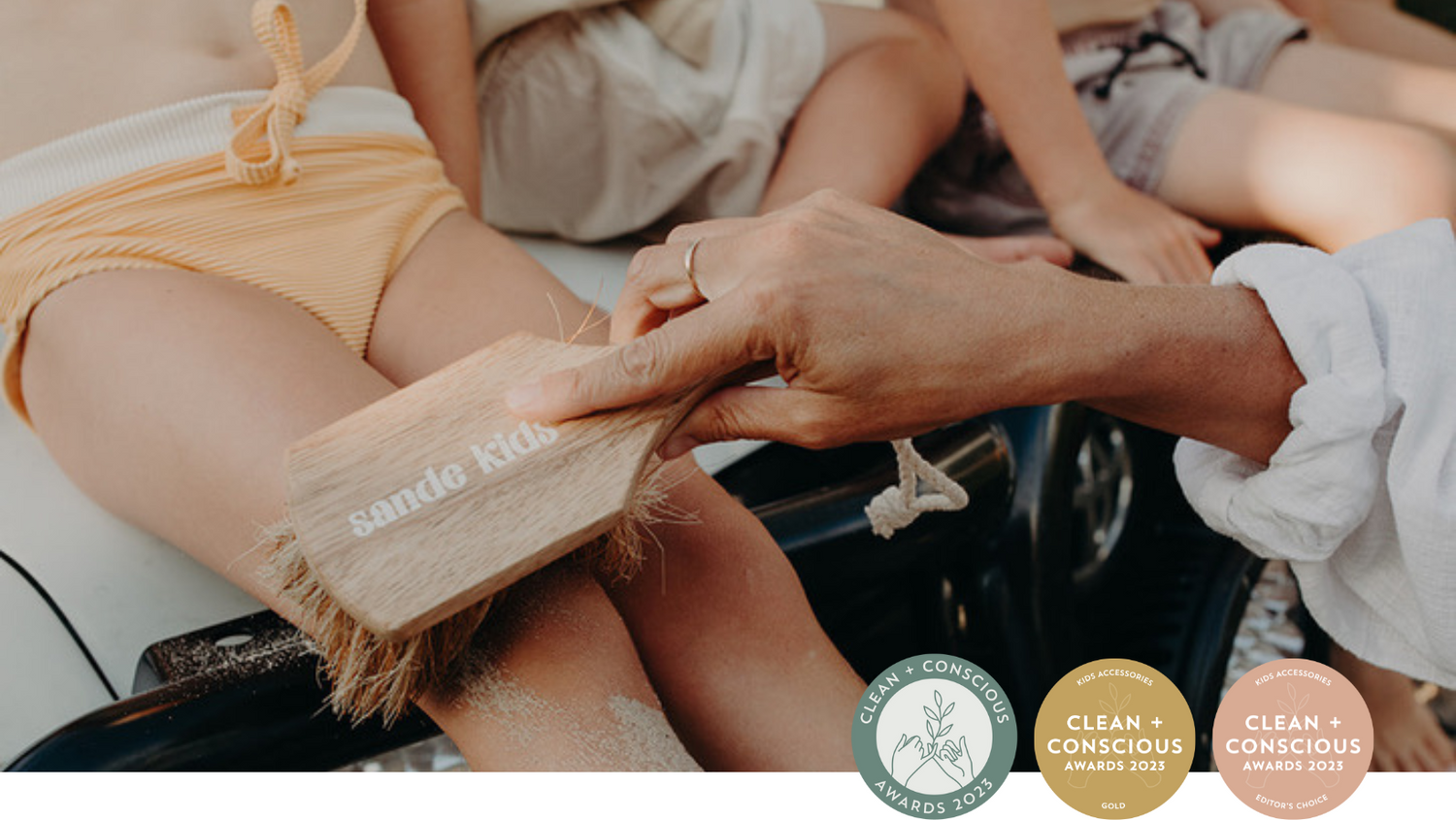 Sande Kids sand brush showing sand being swept off a little girl sitting on the bonnet of a car. Award rosettes overlaid showing Gold and Editor's Choice award from the Clean and Conscious Awards 2023.