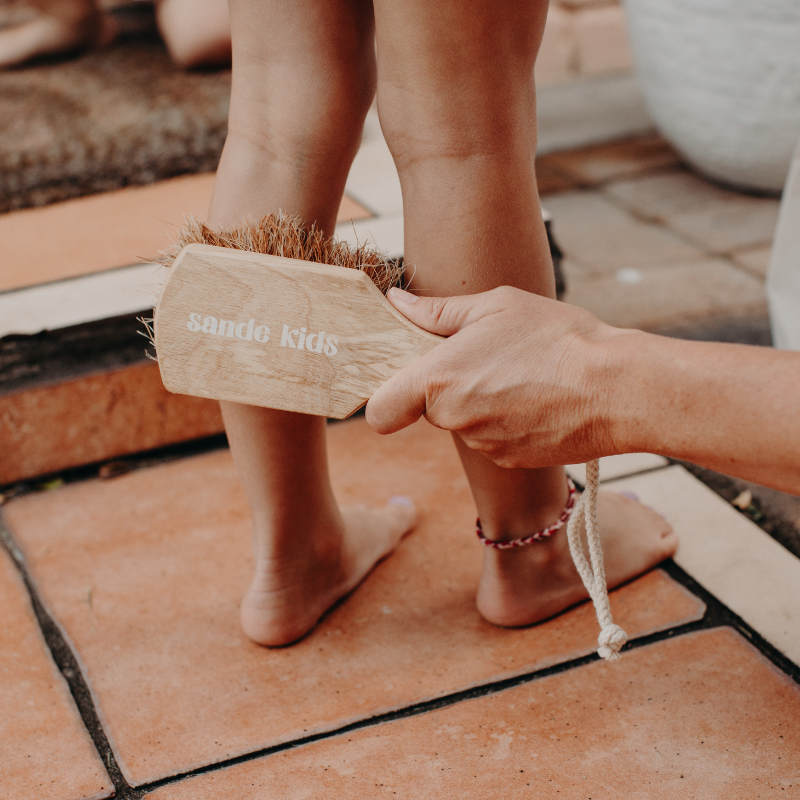 A lifestyle image of a little girl standing on terracotta stairs at the front door, with her legs being swept of sand. The Sande Kids™ Sand Brush is used to gently remove the sand after a day at the beach.