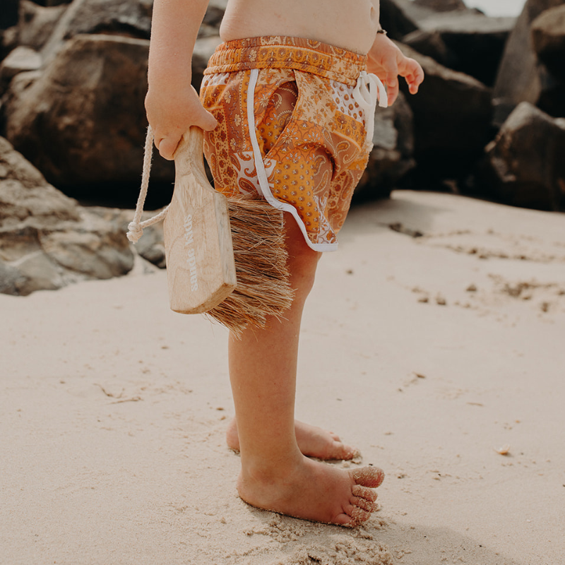 A little boy standing on the beach, holding the Sande Kids™ Sand Brush. Wearing orange board shorts, he’s ready to sweep sand off his feet, with black rocks in the background.