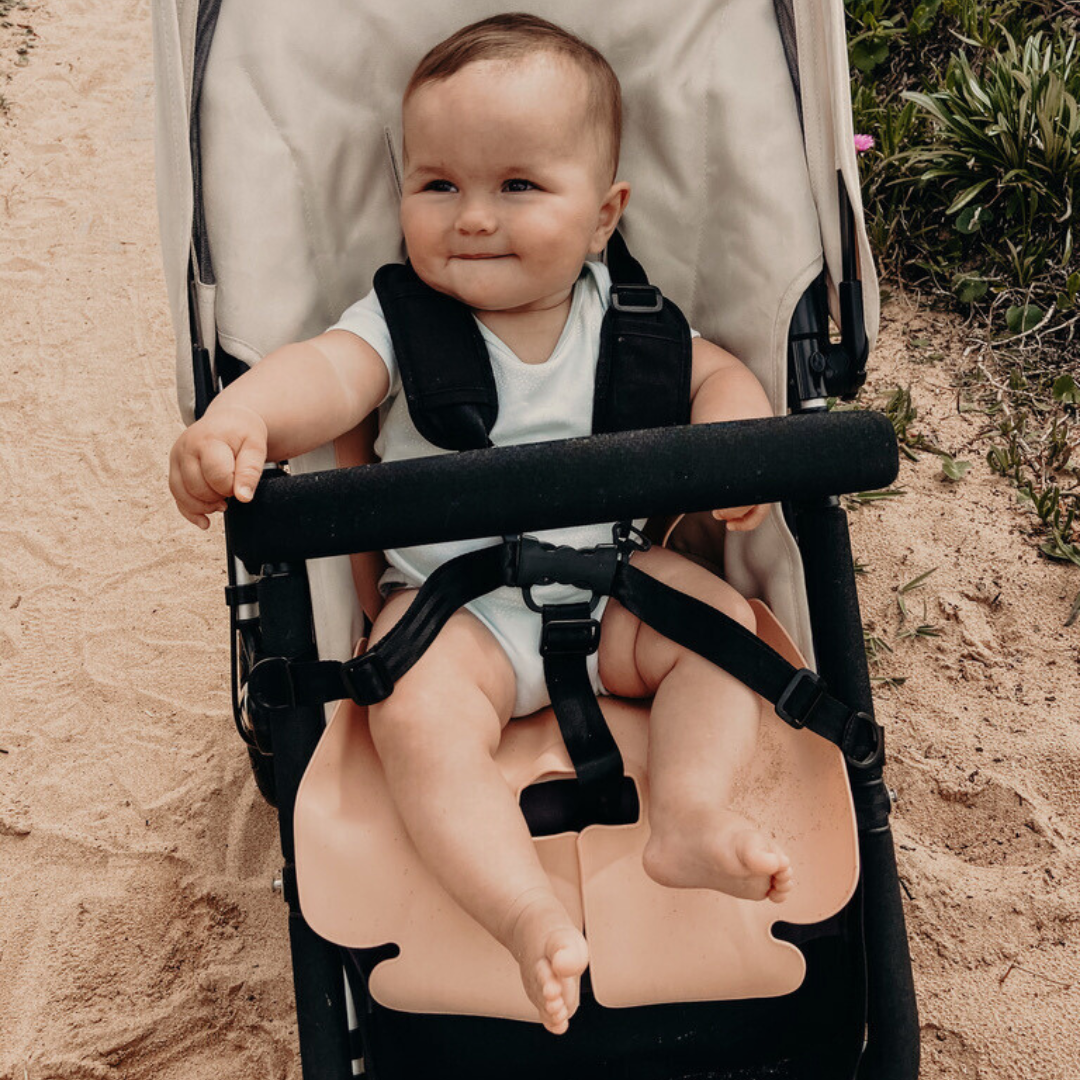 A happy baby girl is sitting in her Bugaboo pram at the beach, resting on the Sande Kids™ waterproof car seat and pram liner in Coral Pink, made from food-grade silicone. Designed for safety and comfort.