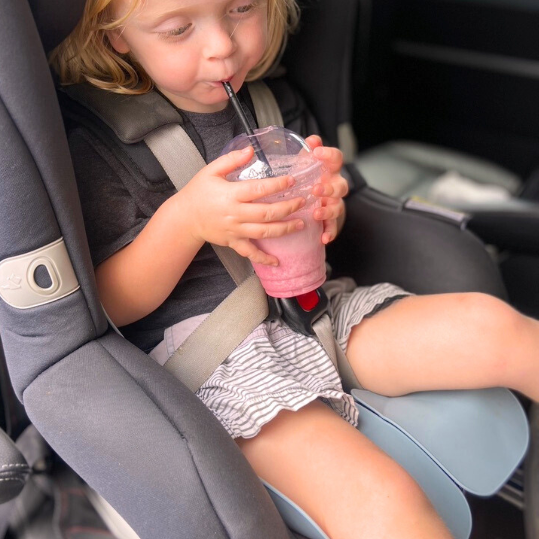 A little boy is sitting in his harnessed car seat, drinking a smoothie from a café. He is seated on the Sande Kids™ waterproof car seat and pram liner in Ocean Blue, which helps avoid sticky messes if anything spills.