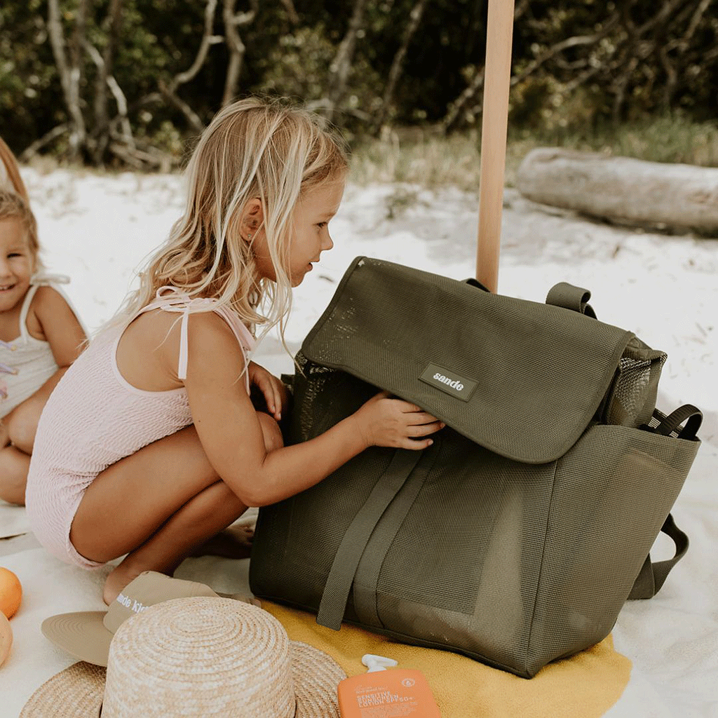 A lifestyle image of a young girl crouching next to the Sande Kids™ Beach Hauler mesh beach backpack in Pandanus Green. The spacious, sand-free mesh backpack features large side pockets for water bottles and toys, adjustable straps, and a durable construction. Ideal for family beach days, outdoor adventures, and carrying kids’ beach essentials while keeping sand out.