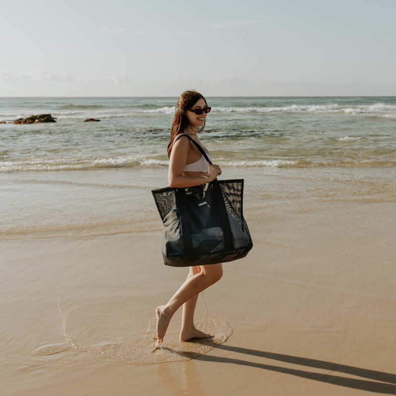 A young woman carrying the Sande™ Beach Hauler - Mini mesh beach bag in Deep Sea Navy along the shoreline at the beach. The transparent mesh reveals beach towels and other essentials inside the bag.