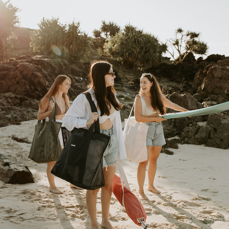 Three friends at the beach carrying Sande Kids Beach Hauler Mini mesh beach bags in Neutral, Olive Green, and Dark Navy. One holds a surfboard, another a beach umbrella, with sand, black rocks, and pandanus palms in the background, showcasing the stylish, spacious, and functional beach bags for friends and family outings.