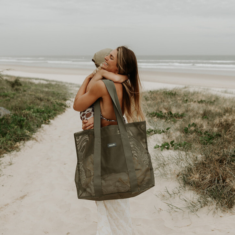 Mother at the beach carrying her toddler son, with a Sande Kids Beach Hauler Mini in Pandanus green on her shoulder, showcasing its convenient size and family-friendly design for beach outings.