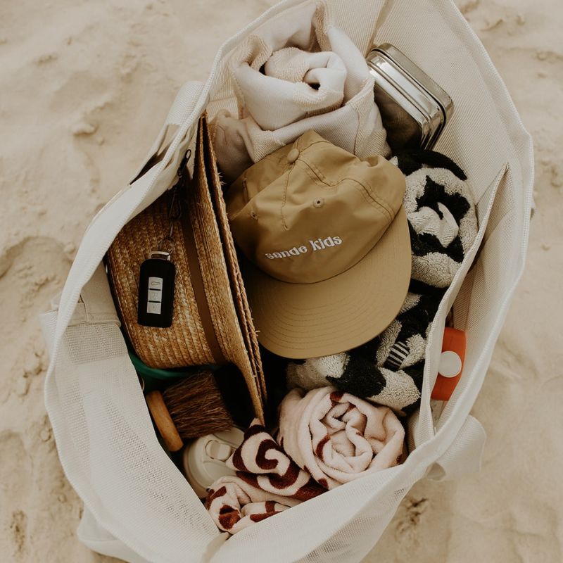 The Sande Kids Beach Hauler mesh beach bag in Sand, resting on the sand - packed with all the essentials for a day at the beach. Showcases the spacious capacity of this oversized beach bag, highlighting how it easily fits beach towels, sunscreen, hats, snacks, water bottles, and valuables—making it the perfect beach bag for all your essentials.