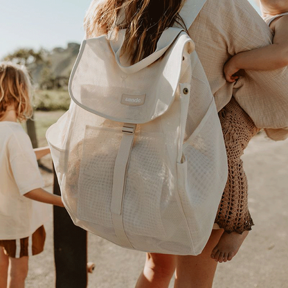 Lifestyle image of a mum carrying a toddler on her hip with the Sande Kids Beach Hauler Mesh Backpack in Sand on her back, ready for a beach day. Features spacious design and adjustable straps for comfortable, hands-free carrying of beach essentials.