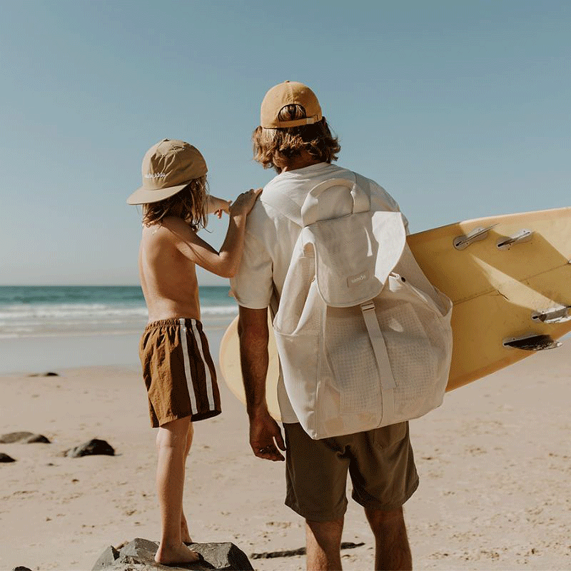 Dad and son checking the surf at the beach, with dad carrying a yellow surfboard and the Sande Kids Beach Hauler Mesh Backpack, while the son wears a Sande Kids branded cap, highlighting the backpack’s spacious, durable design for beach and surf outings.