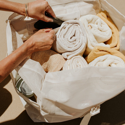 A close-up of a phone being placed into the secure pocket of the Sande Beach Hauler™ Backpack, packed with rolled towels, a hat and drink bottles to show what fits inside the oversized neutral mesh beach backpack.