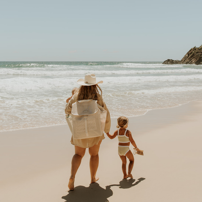 A woman and young girl walk hand in hand along the beach. The woman carries a cream Sande™ Beach Hauler Mesh Backpack and wears a straw hat and beige cover-up.