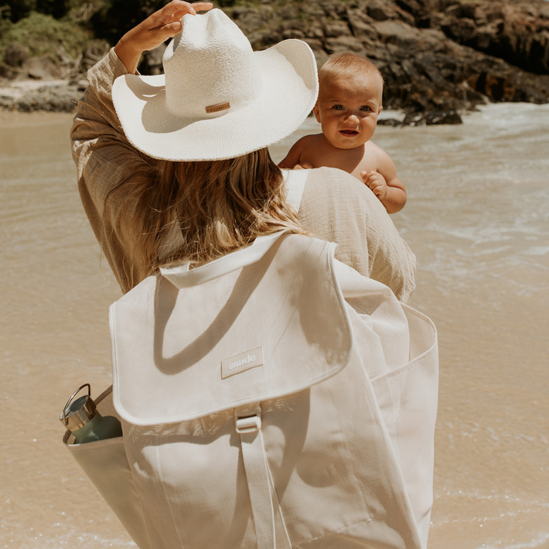 Lifestyle image of a mum carrying a cute baby at the beach with the Sande™ Beach Hauler Mesh Backpack in Sand on her back, ready for a beach day. Features spacious design and adjustable straps for comfortable, hands-free carrying of beach essentials.