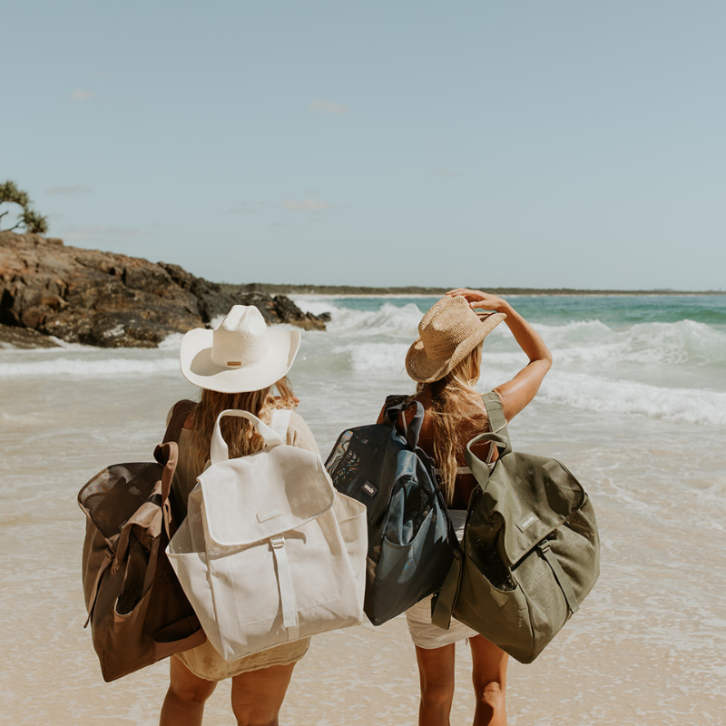 Two women standing on a beach from behind, each carrying 2 Sande Beach Hauler mesh backpacks in different colours: one brown, one cream, one nave and one olive green. They are wearing wide-brimmed hats and light clothing, with the ocean and clear blue sky in the background.