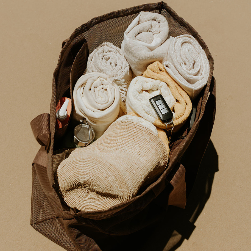 Top view of the Sande oversized mesh beach bag showing capacity with six rolled towels, a sand brush, sunscreen in the internal mesh pocket, and car keys clipped to the key holder. The bag sits on the sand.