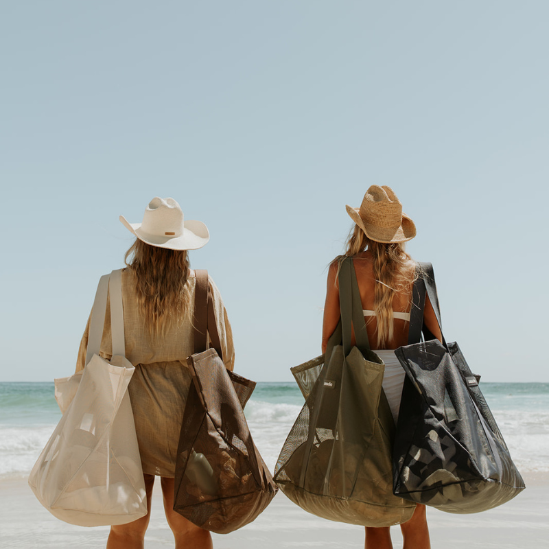Two women standing on a beach from behind, each carrying 2 Sande Beach Hauler mesh beach bags in different colours: one cream, one brown, one olive green and one deep navy. They are wearing wide-brimmed hats and light clothing, with the ocean and clear blue sky in the background.
