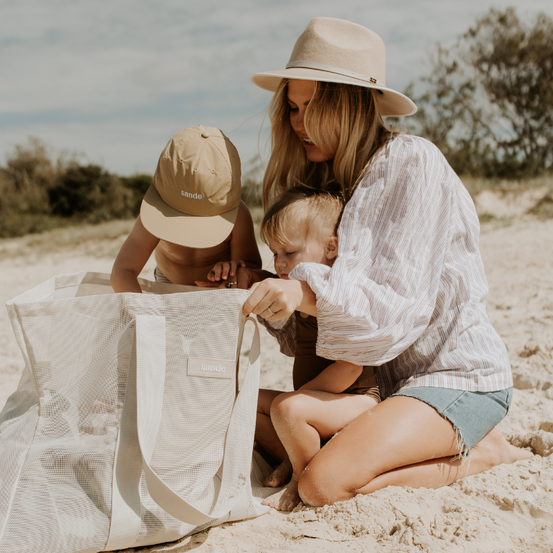 Lifestyle image of a young family looking into their neutral Sande™ mesh beach bag. A little boy wearing a Sande™ cap reaches into the bag, while a little girl sits on her mothers lap. The mother has long blonde hair and is wearing a white shirt, denim shorts, and a cream fedora hat