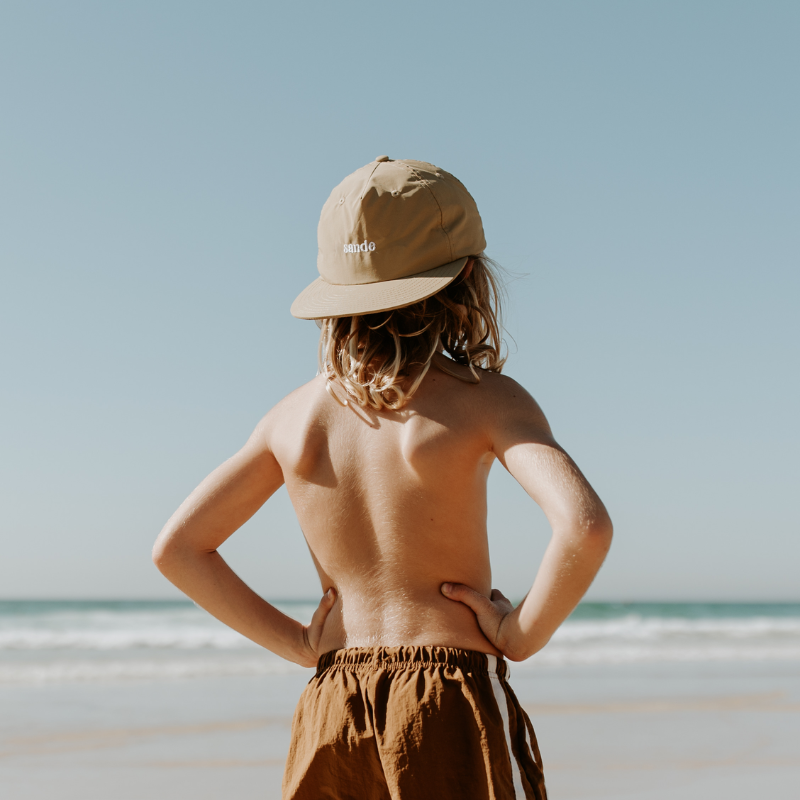 A lifestyle image of a young boy with the Sande™ cap worn backwards. He stands on the beach, hands on hip, looking out to the ocean.