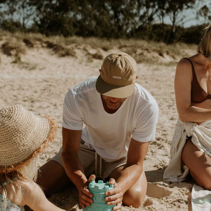 A lifestyle image of dad wearing the Sande™ cap while building sandcastles on the beach with his family.