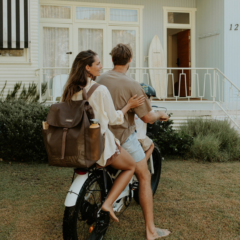 Couple riding a e-bike in front of a house with a surfboard in the background. the women on the back wears the Sande Beach Hauler Mesh Backpack in Espresso chocolate brown, showcasing its versatile, hands-free design for active beach commutes and outdoor adventures.