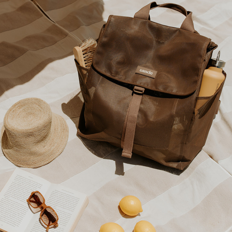 Lifestyle image of the Sande Beach Hauler Mesh Backpack in Espresso chocolate brown, showcasing its spacious design and durable construction for carrying family beach essentials. The backpack sits on a neutral towel with a book, sunglasses, a hat, and fresh lemons nearby. A sand brush and drink bottle are visible in the side pockets of the fully closed backpack.