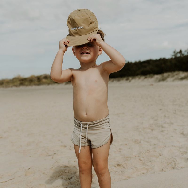 A lifestyle image of a little boy holding the Sande Kids™ cap just above his head, with a smile on his face. He stands on the beach, with vegetation and dunes in the background.