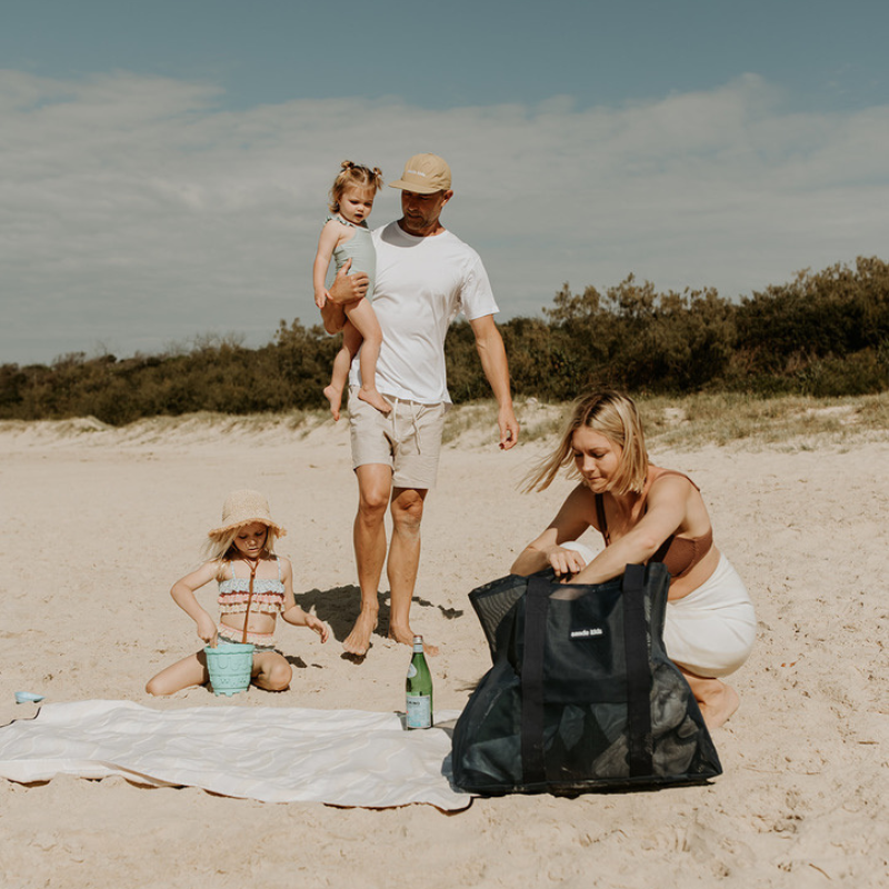An image of a family at the beach, with a little girl playing with a bucket. Dad is wearing a Sande Kids Cap and carrying a little girl. A beach towel is laid out. The mum, crouching down next to her Deep Sea Navy Beach Hauler mesh beach bag. The beach setting is complemented by grassy dunes and vegetation in the background.