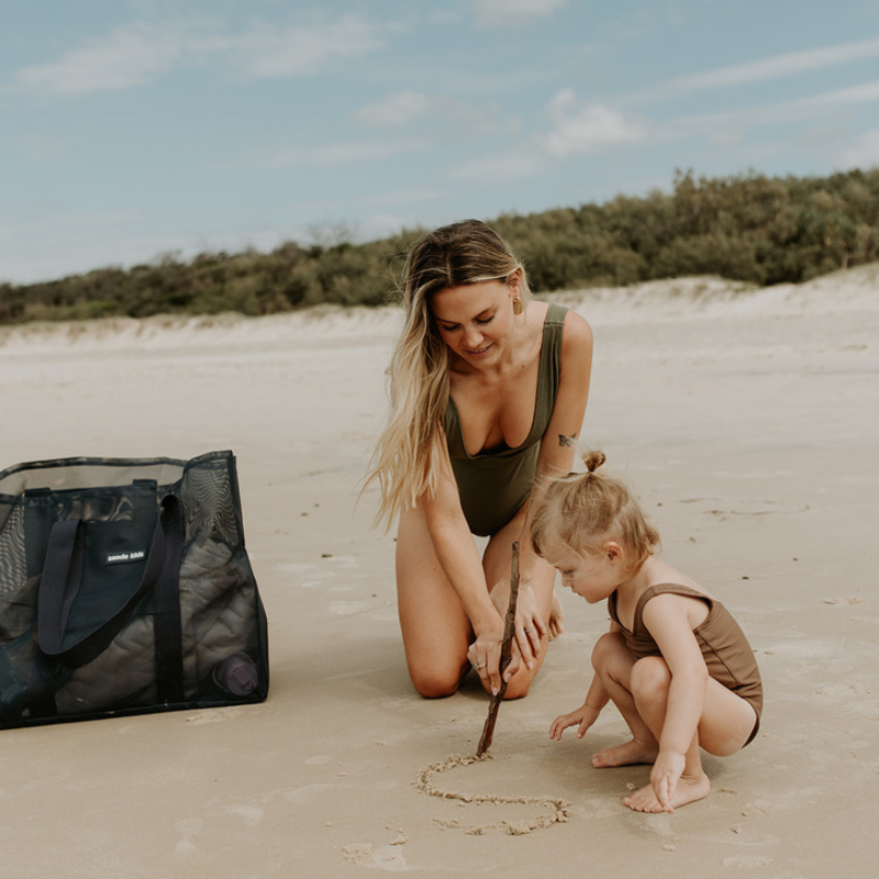 A mum and little girl at the beach, drawing with a stick in the sand. Next to them is their Sande Kids Beach Hauler mesh beach bag in Deep Sea Navy, resting on the sand.