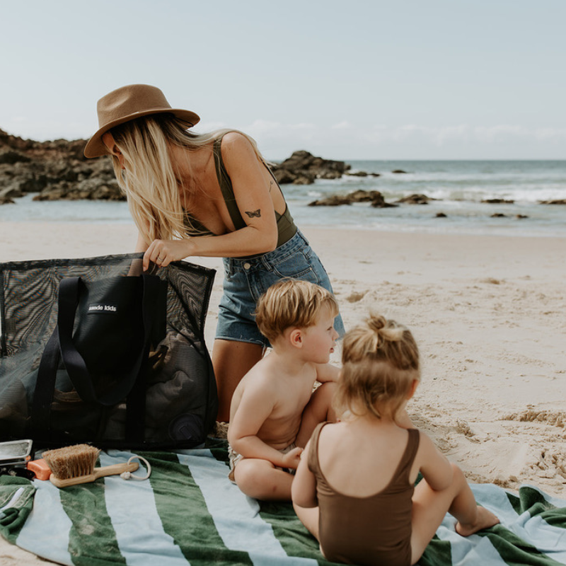 A mum and two little kids at the beach, sitting on a green and blue striped beach towel. The mum is rummaging through her oversized mesh beach bag by Sande Kids, the Beach Hauler mesh bag in Deep Sea Navy. The ocean is visible in the background.