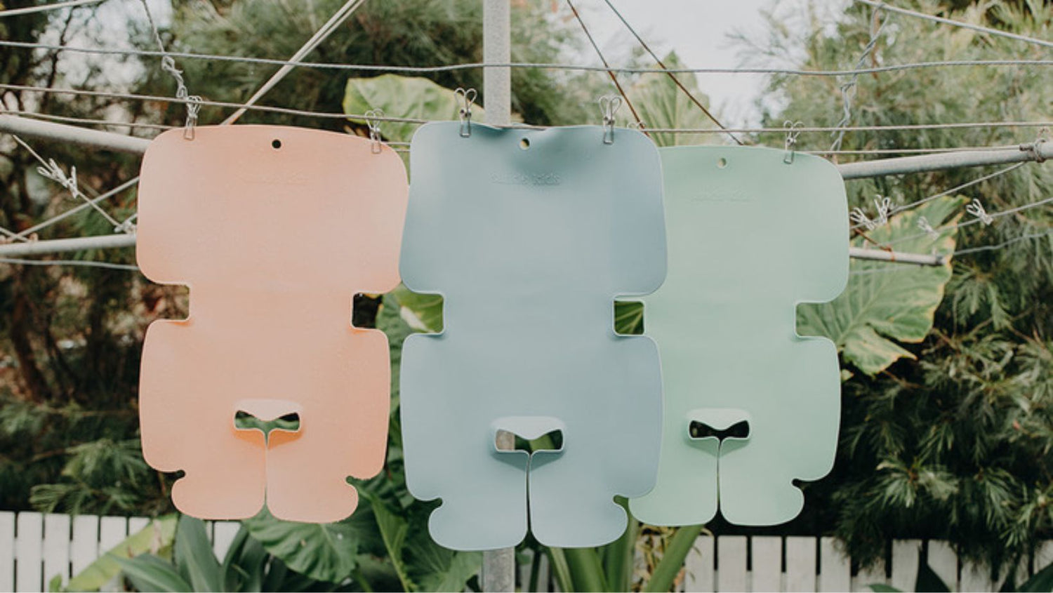 Sande Kids children's waterproof car seat and pram liners made from food grade silicone hanging on a hillshiost clothesline. Showing colours; Coral pink, Ocean blue and Seafoam green.