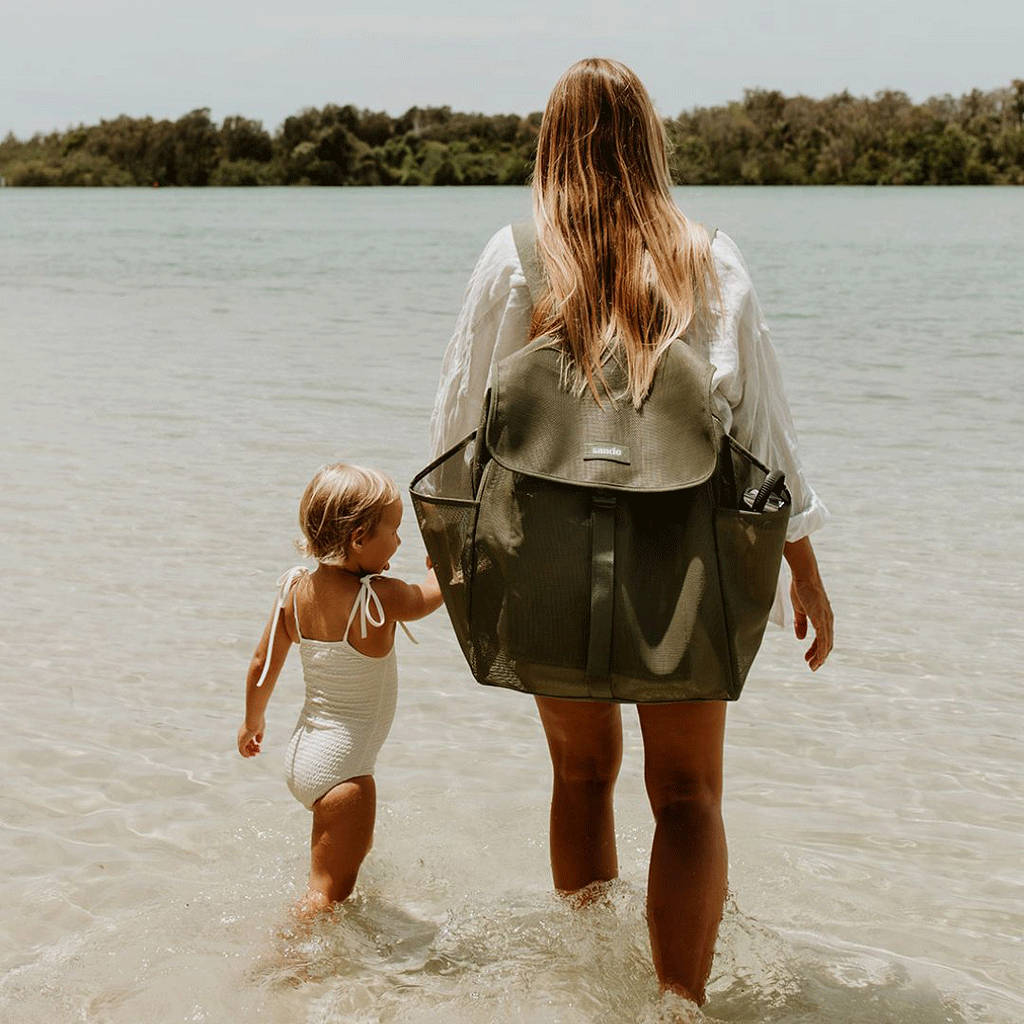A woman with long blonde hair stands at the water’s edge, holding her little girl’s hand. She wears a dark olive green Sande Kids™ Beach Hauler mesh backpack, which features side pockets, a flap with a clip, and a white silicone logo. The scene captures a peaceful beach moment, highlighting the backpack’s style and practicality for family outings.