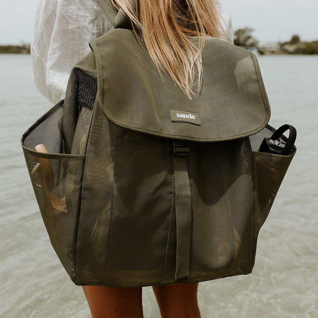 A woman with long blonde hair stands at the water’s edge wearing the Sande Kids™ Beach Hauler mesh beach backpack in Pandanus green. This sand-free mesh beach bag features large capacity storage, breathable mesh fabric, side pockets for water bottles, and a secure clip-up flap. The lightweight family beach backpack displays a silicone Sande Kids™ logo and is designed for parents, beach days, travel, and carrying kids’ beach gear while keeping sand out.
