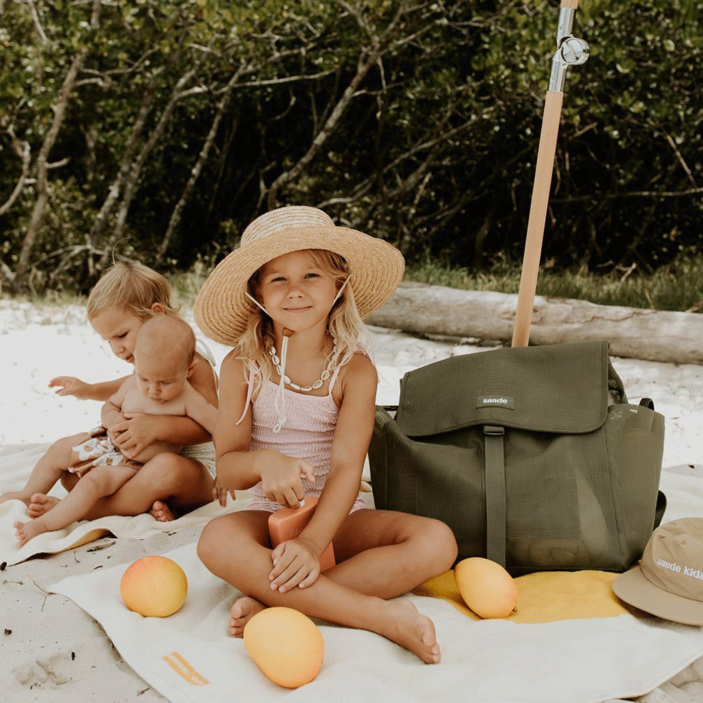 A lifestyle image of young girls sitting under a beach umbrella surrounded by beach towels, sunscreen, and mangoes. One girl wears a wide-brim straw hat and sits next to the Sande Kids™ Beach Hauler mesh beach backpack in Pandanus Green. The spacious, sand-free backpack features large side mesh pockets for water bottles and toys, adjustable straps, and a durable construction. Perfect for family beach days, outdoor adventures, and carrying kids’ essentials while keeping sand out.