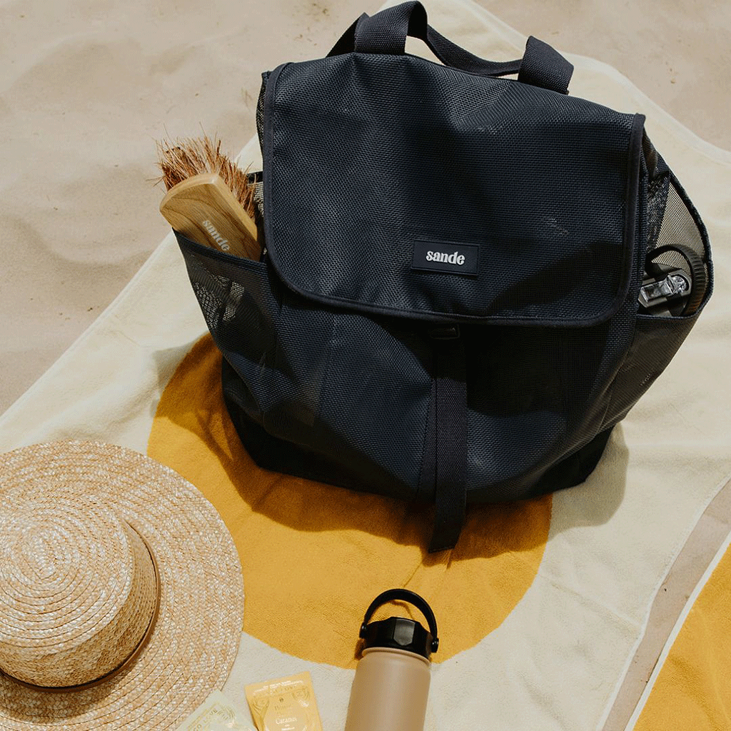 Top view of the Sande Kids™ Beach Hauler mesh backpack, closed and sitting on a beach towel on the sand. A Pargo drink bottle and a Sande Kids™ brush are placed in the outer mesh pockets.