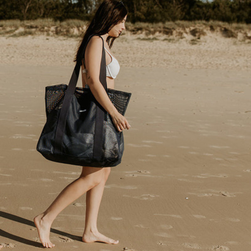 A young woman walking along the beach carrying the Sande Kids Beach Hauler - Mini mesh beach bag in Deep Sea Navy. 