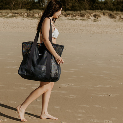 A young woman walking along the beach carrying the Sande Kids Beach Hauler - Mini mesh beach bag in Deep Sea Navy. 
