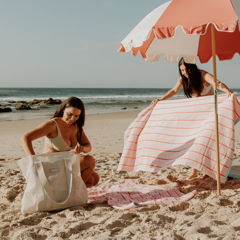 Friends enjoying a day at the beach with their Sande™ Beach Hauler Mini mesh bags in a neutral tone. Pink and red striped beach towels and a coral red and white beach umbrella create a vibrant, fun setting for a perfect beach day.