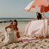 Friends enjoying a day at the beach with their Sande™ Beach Hauler Mini mesh bags in a neutral tone. Pink and red striped beach towels and a coral red and white beach umbrella create a vibrant, fun setting for a perfect beach day.